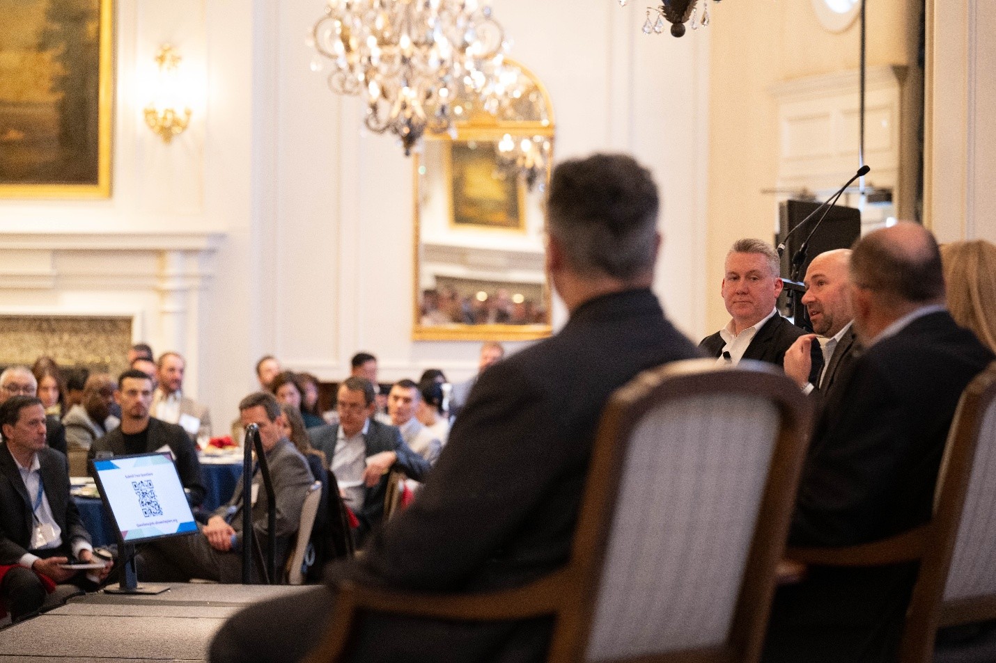 In this photo, from a distance, Jason Martin is facing in the direction of the camera but looking to Steve Wallace sitting to Martin's left. Martin and Wallace are framed by the heads of two other panelists sitting with them on a stage. To the lower left of the panelists there are several attendees looking to the panel. The room has high ceilings a large fireplace on the left of the image with a large framed painting. In the center of the image is a large framed mirror, and an ornate chandelier hangs from the ceiling.